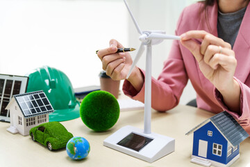 A businesswoman in a pink suit stands at a white desk with a world model, electric car model, and green light bulb, promoting Earth Day, green energy, and sustainability efforts.