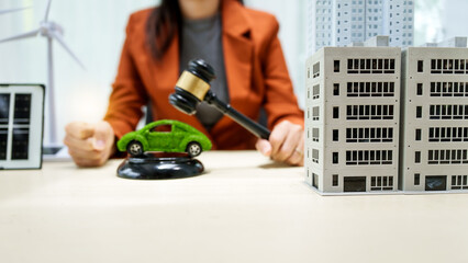 A young businesswoman is seated at a white desk, holding a lawyer's gavel. The city buildings and an electric car symbolize a commitment to green energy and sustainable policies, green car toy model.