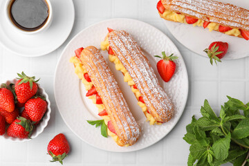 Delicious eclairs filled with cream, strawberries, mint and tea on white tiled table, flat lay