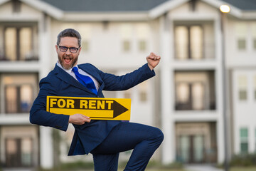 Realtor agent is a realtor with sign for rent in hand against the background on new apartment home background. Realtor in suit, outdoor portrait. Realtor renting new home. Property rental concept.