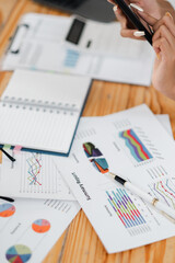 Close-up of hands holding a smartphone over financial charts, graphs, and reports on a wooden desk, showcasing business analysis and planning.