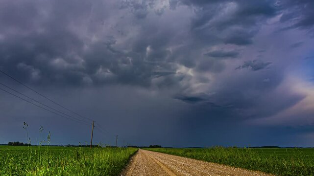 Dramatic dirt road between field of flowers against sky with black clouds in timelapse. Agricultural Landscape. Time-lapse of storm clouds
