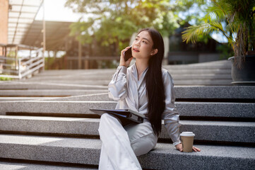 Beautiful Asian businesswoman is talking on the phone outdoors on the stairs during her coffee break