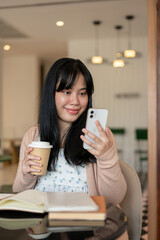 Fototapeta premium A happy Asian woman relaxing in a coffee shop, having a hot drink while using her smartphone.