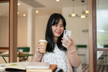 A happy Asian woman relaxing in a coffee shop, having a hot drink while using her smartphone.