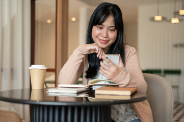 A happy, beautiful Asian woman using her smartphone at a table indoor while reading books.