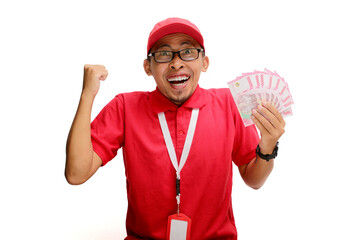 Excited Asian delivery man or courier raises his fist in triumphant YES gesture while holding money banknotes in his hand, isolated on white background. Concepts of victory, financial gains, triumphs
