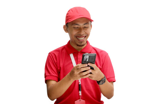 A cheerful Asian delivery man or courier, wearing a red cap and red polo shirt, texting on his phone, receiving good news about a successful transaction or delivery. Isolated on a white background