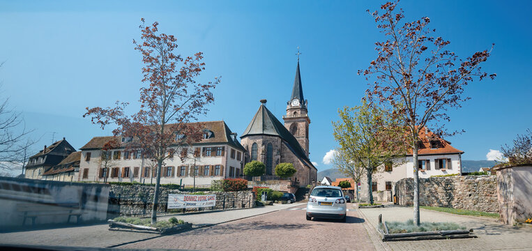 Bergheim, France - Apr 19, 2019: A charming street in a French village featuring a stone church with a tall spire, residential buildings, and a parked car, with a banner reading DONNEZ VOTRE SANGky
