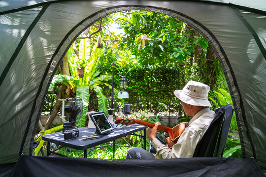 Senior man sitting and playing guitar With a table and equipment for morning coffee. The atmosphere of setting up a tent and camping in the forest.