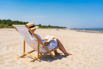 Summer holidays on the Baltic Sea. Woman of European beauty in white tunic and beige hat reading  book sitting on deckchair on beach with white sand on beautiful sunny day. Relaxing on beach