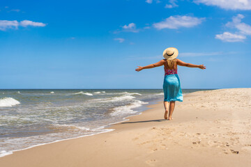 Summer vacation. Beautiful young woman in colorful swimsuit and blue pareo and hat walking with her arms spread out on the sandy beach on a beautiful sunny day. Back view