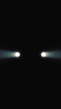 Front view of turning on the headlights in the car at night. Media. Abstract white lights of a car on a black background. Vertical video.