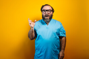 A man in a blue blouse and glasses stands in front of a yellow background, with his right hand...