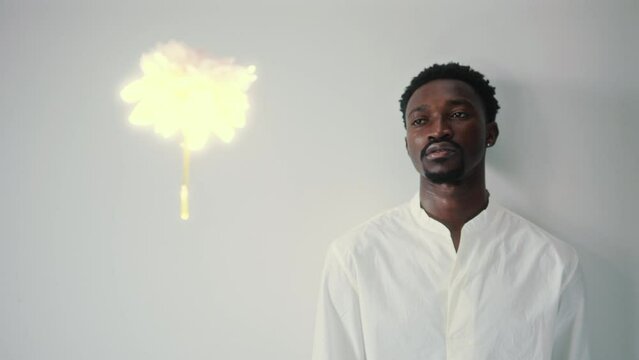 Young relaxed Black man leaning back on the wall and looking at white glowing flower levitating and spinning in the air. Studio shot, rack focus