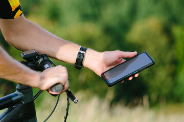 Cyclist man holding a white screen smartphone mockup on the background of a bicycle in nature. Hand with smartphone close-up