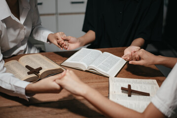 Group of Christians sit together and pray around a wooden table with blurred open Bible pages in their homeroom.