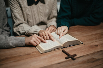 Group of Christians sit together and pray around a wooden table with blurred open Bible pages in their homeroom.