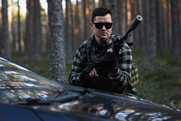 A man in sunglasses is a private military man with a rifle with a silencer near a car in the forest.