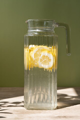 Jug with lemonade drink on wooden table with natural floral sunlight shadows, soft green background, lifestyle, healthy summer homemade beverage