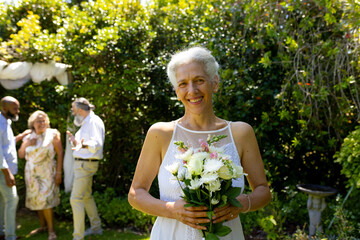 Smiling senior bride holding bouquet of flowers at outdoor wedding with friends