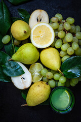 Flat lay of grapes, lemons, pears and glass of lemon juice with green leaves, dark background, vertical