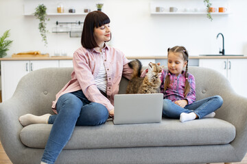 Family moment with mother and daughter sitting on couch using laptop with cat. Cozy home setting in modern kitchen. Bonding time, pet-friendly environment, technology use