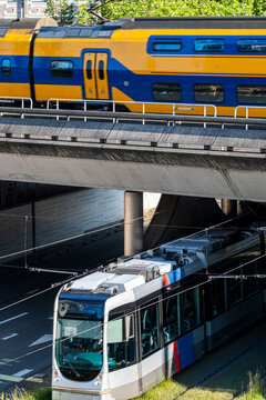 A yellow and blue train crosses an elevated overpass while a city tram navigates below on the street level, showcasing a multi-layered urban transport system.