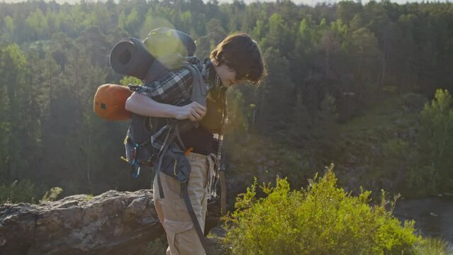 Lens flared tilt up full shot of young woman putting on heavy hiking backpack on her shoulders and continuing her summer journey