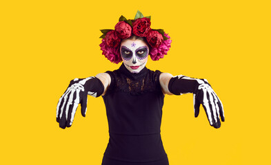 Portrait of woman in Halloween or Day of the Dead costume. Young lady in black dress, skeleton gloves, flower wreath and Mexican Catrina sugar skull mask make up standing isolated on yellow background