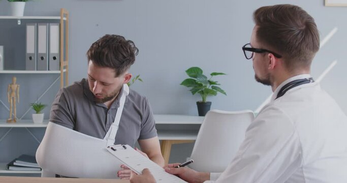 Patient with a hand injury seeing a doctor at the clinic. Doctor with a clipboard sitting at his desk in the office talking to a man complaining about pain and discomfort while wearing an arm sling