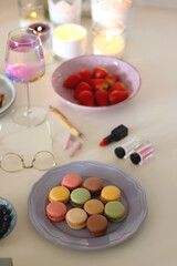 Plate of pastel macarons, cookies and chocolate, cup of tea of coffee, glass of bubble water, various berries, books and accessories on the table. Selective focus, pastel colors.