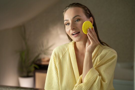 A woman in a yellow robe applies facial cleanser with a sponge in the bathroom. She gently rubs the cleanser on her face in a circular motion