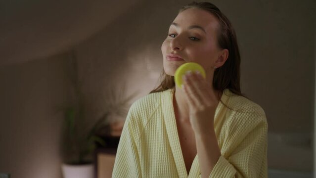 A woman in a yellow robe applies facial cleanser with a sponge in the bathroom. She gently rubs the cleanser on her face in a circular motion