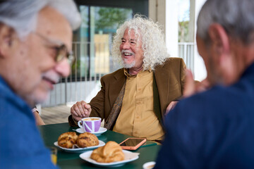 Mature grey haired man smiling enjoying coffee with his group of friends on terrace of old people home. Meeting of senior Caucasian male persons sitting together having snack or breakfast outdoors