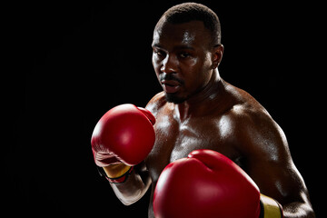 Portrait of determined African-American man, shirtless boxing athlete standing in red gloves,...