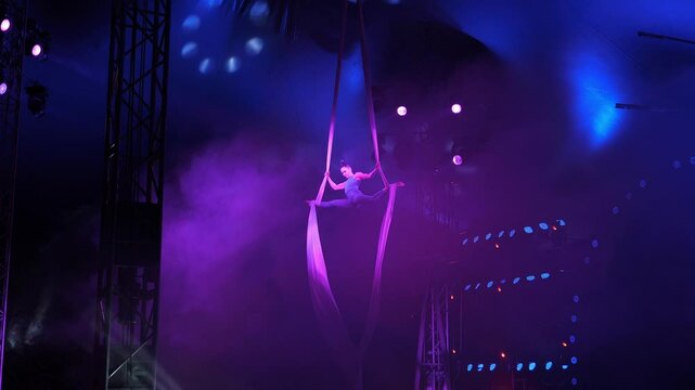 An aerialist performs on a suspended fabric in the circus arena, she performs complex tricks. An acrobat holding her feet to the fabric does the splits under the dome of the circus.
