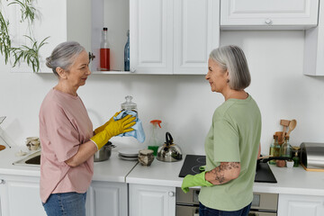 A mature lesbian couple cleans their modern apartment kitchen together. One woman is wearing yellow gloves and wiping a glass jar. - Powered by Adobe