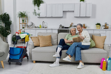 Obraz premium Two mature lesbian women rest on a sofa in their modern apartment after cleaning, with cleaning supplies visible in the background.