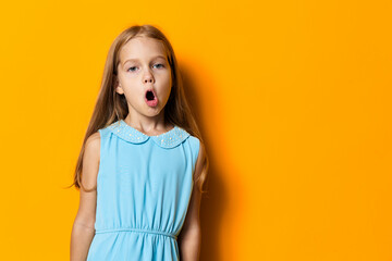 Joyful little girl in blue dress expressing excitement with wide open mouth against vibrant orange...