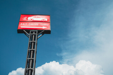 Red drive thru sign with car and arrow symbols, standing high against a blue sky background with white puffy clouds.