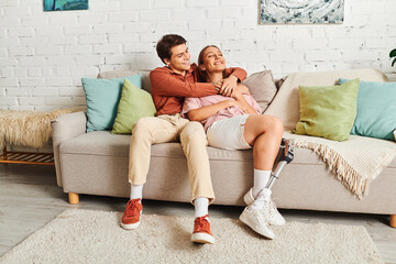 A couple sits on a couch, the woman wearing a prosthetic leg, with her boyfriend holding her close.