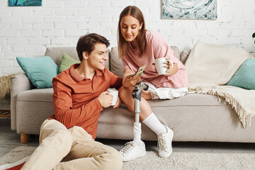 Woman with prosthetic leg sits on couch with boyfriend, sharing coffee and intimate moment.