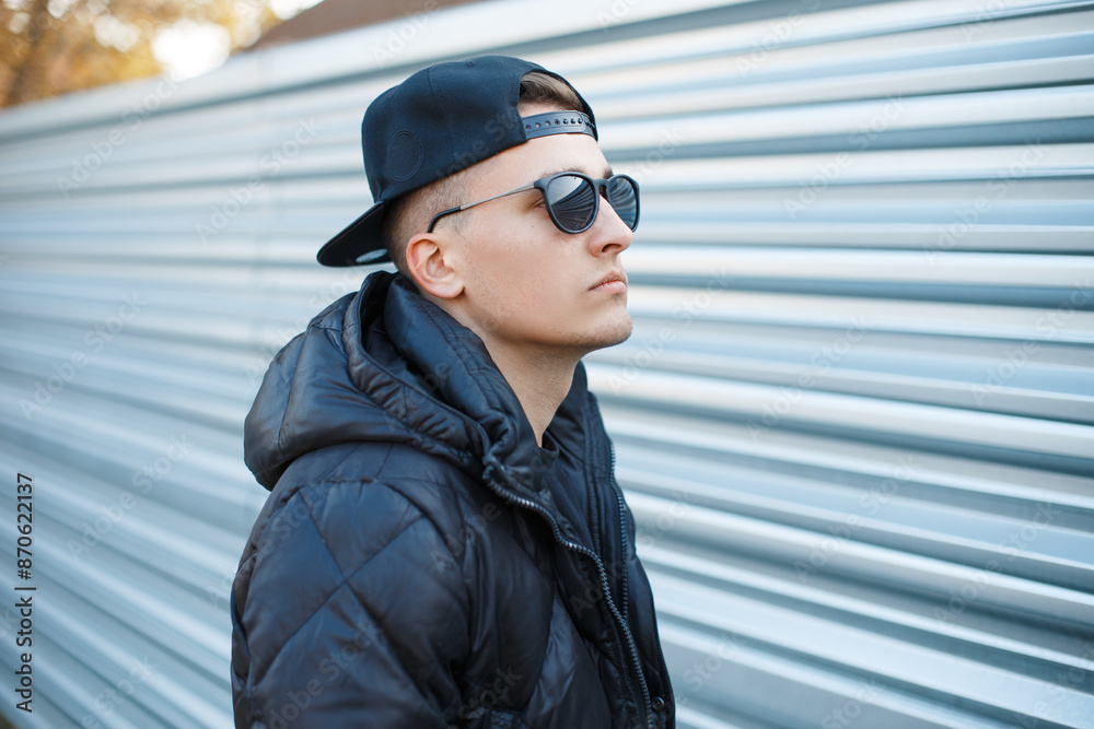 Poster Handsome fashion handsome hipster man with a baseball cap with cool sunglasses in a fashionable black down jacket on the street on a metal background