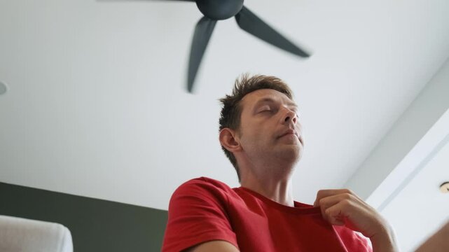 Man sitting under ceiling fan at home in living room, cooling off during hot summer weather, suffering from heat, high temperature. Sweaty exhausted male lifting his red shirt to get cool