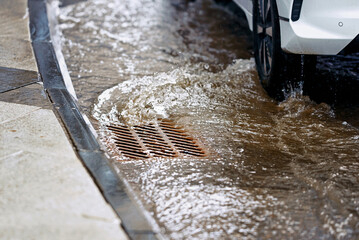 Flood water rushing over road and into storm drain grate, car driving along flooded road during...