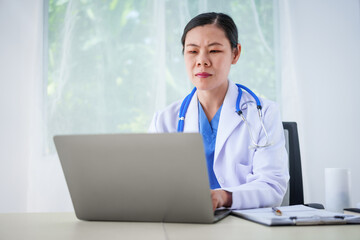 An Asian female doctor sits at her desk, conducting an online consultation. This faster, cheaper, and more convenient method helps prevent disease spread and offers easier access to healthcare.