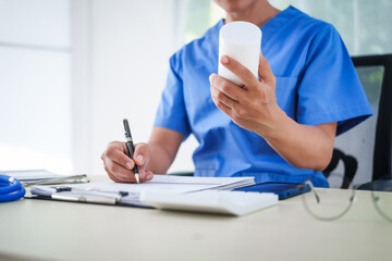 Fototapeta premium An Asian female nurse sits at a table, talking on the phone with a patient, explaining medicine during an online consultation. This faster, cheaper method helps prevent disease spread and eases