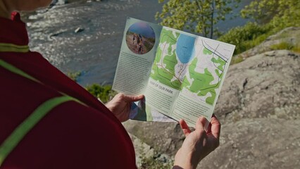 Over shoulder of unrecognizable tourist standing by river in sunlight and reading brochure of national park with map while on summer solo trip in nature