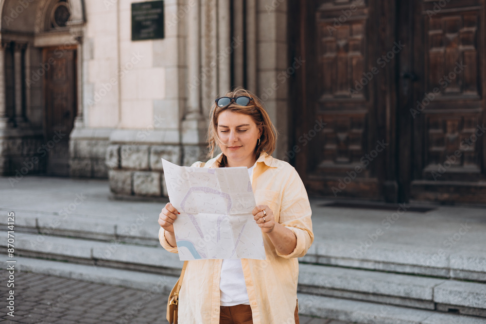 Wall mural Happy young woman with map near old door in city. Holidays and tourism concept. Female tourist searching direction on location map while traveling abroad in summer